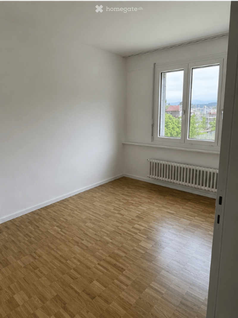 Empty room with wooden flooring, white walls, and a window with a radiator below it.