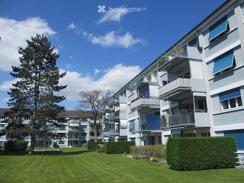 Multi-story residential building with balconies, surrounded by a well-maintained lawn and trees.