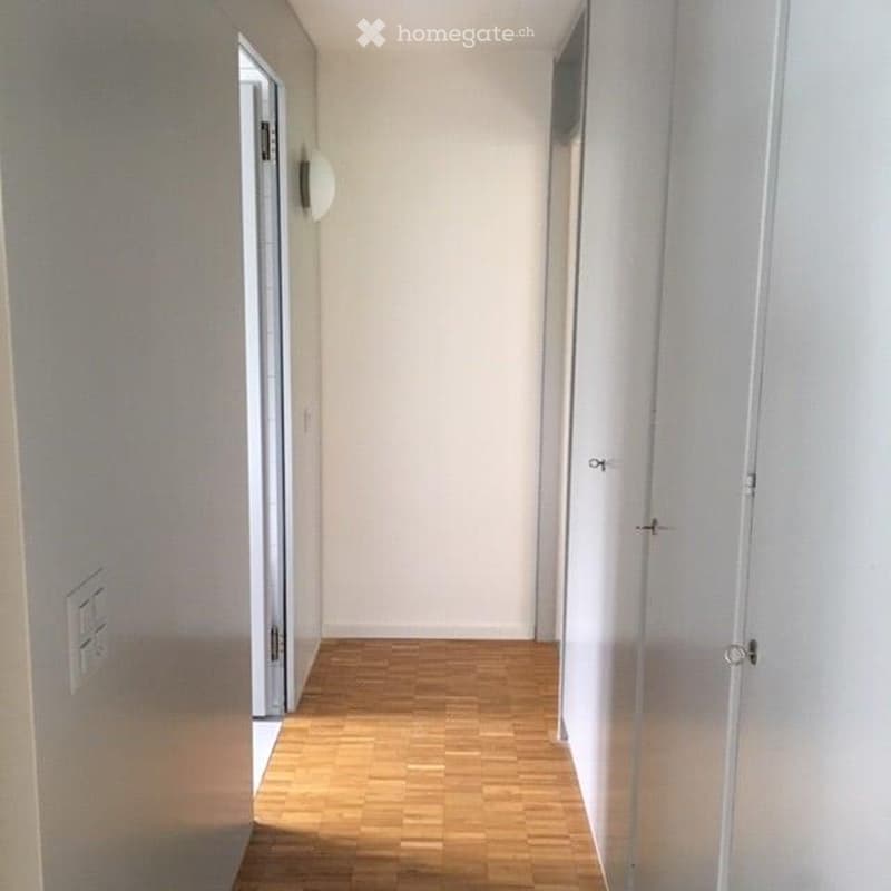 A narrow, well-lit hallway with light wooden flooring and white walls. There are two doors on either side, one with a visible handle, and a small wall-mounted light fixture on the left side.