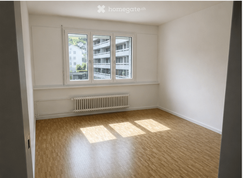 Empty room with wooden flooring, white walls, and large windows offering a view of the outside buildings.
