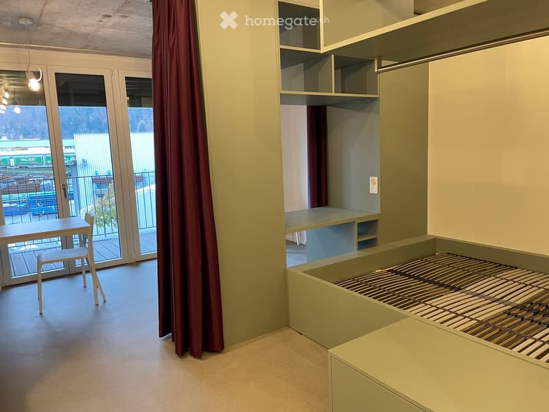 Modern, minimalist room with built-in bed platform, shelves, a small table, and a chair near a large window with a view of the street.