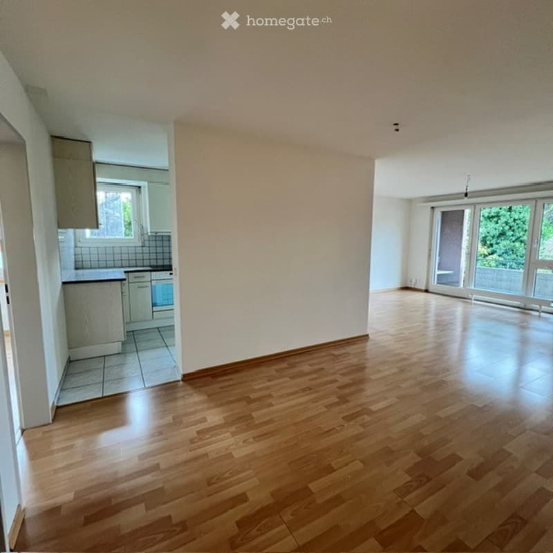 Empty room with wooden flooring, white walls, and large windows leading to a balcony. The adjacent kitchen area is also visible with cabinets and a countertop.
