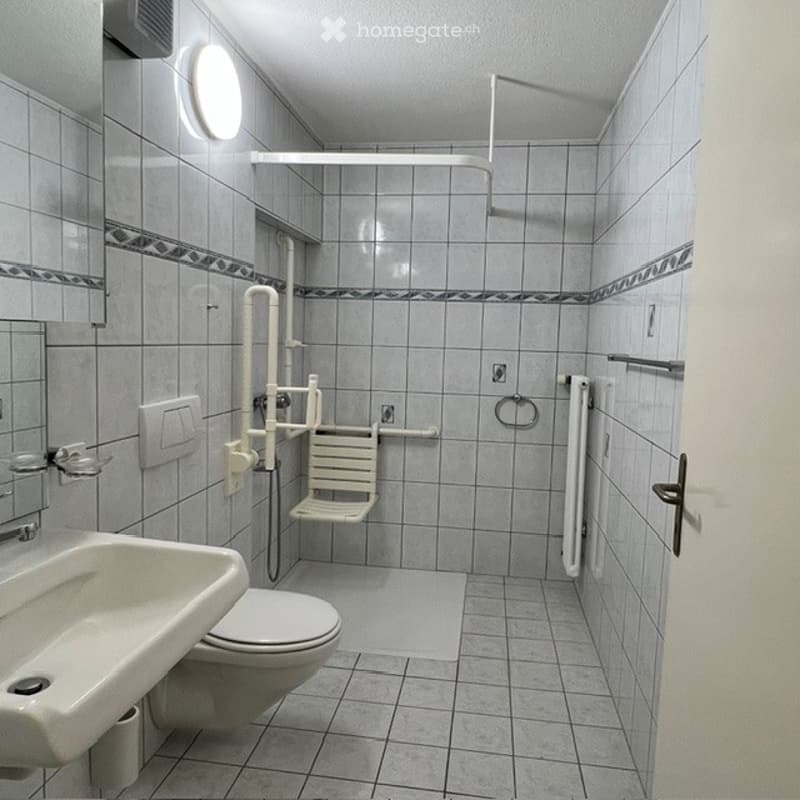 Accessible bathroom with tiled walls and floor, featuring a toilet, sink, shower chair, handrail, and mirror.