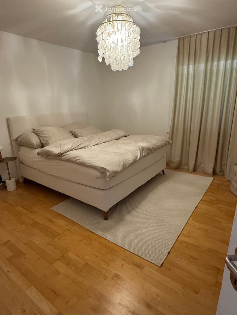 Bedroom with a large bed, nightstand, chandelier, and curtains. Wooden flooring and a small rug.