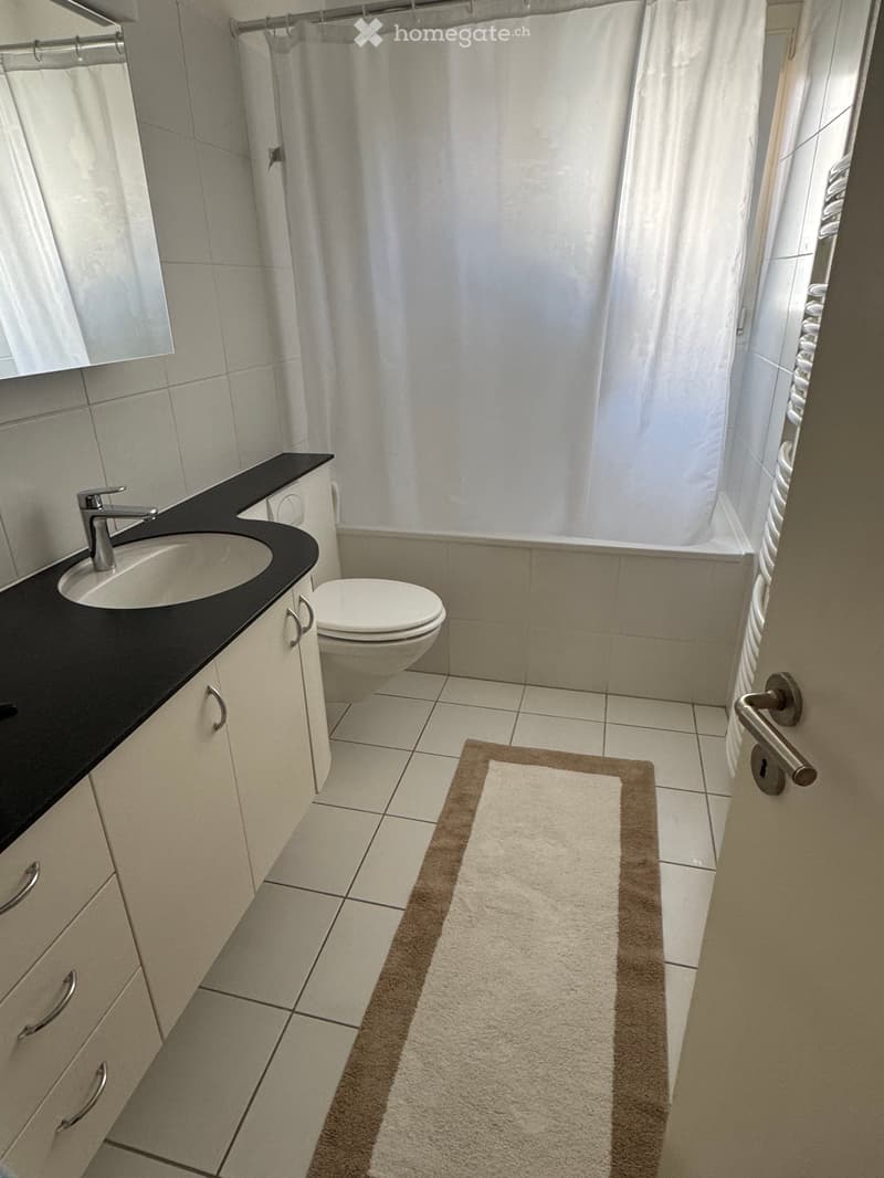 Modern bathroom with a white tiled floor, black countertop, white sink, silver faucet, toilet, and a shower with a white curtain. A beige bath mat is placed in front of the shower area.