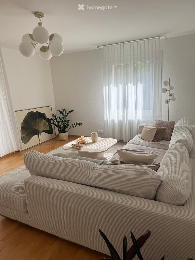 Modern living room with a grey sectional sofa, coffee table, floor lamp, and potted plants. Large windows with sheer curtains allow natural light to fill the space.