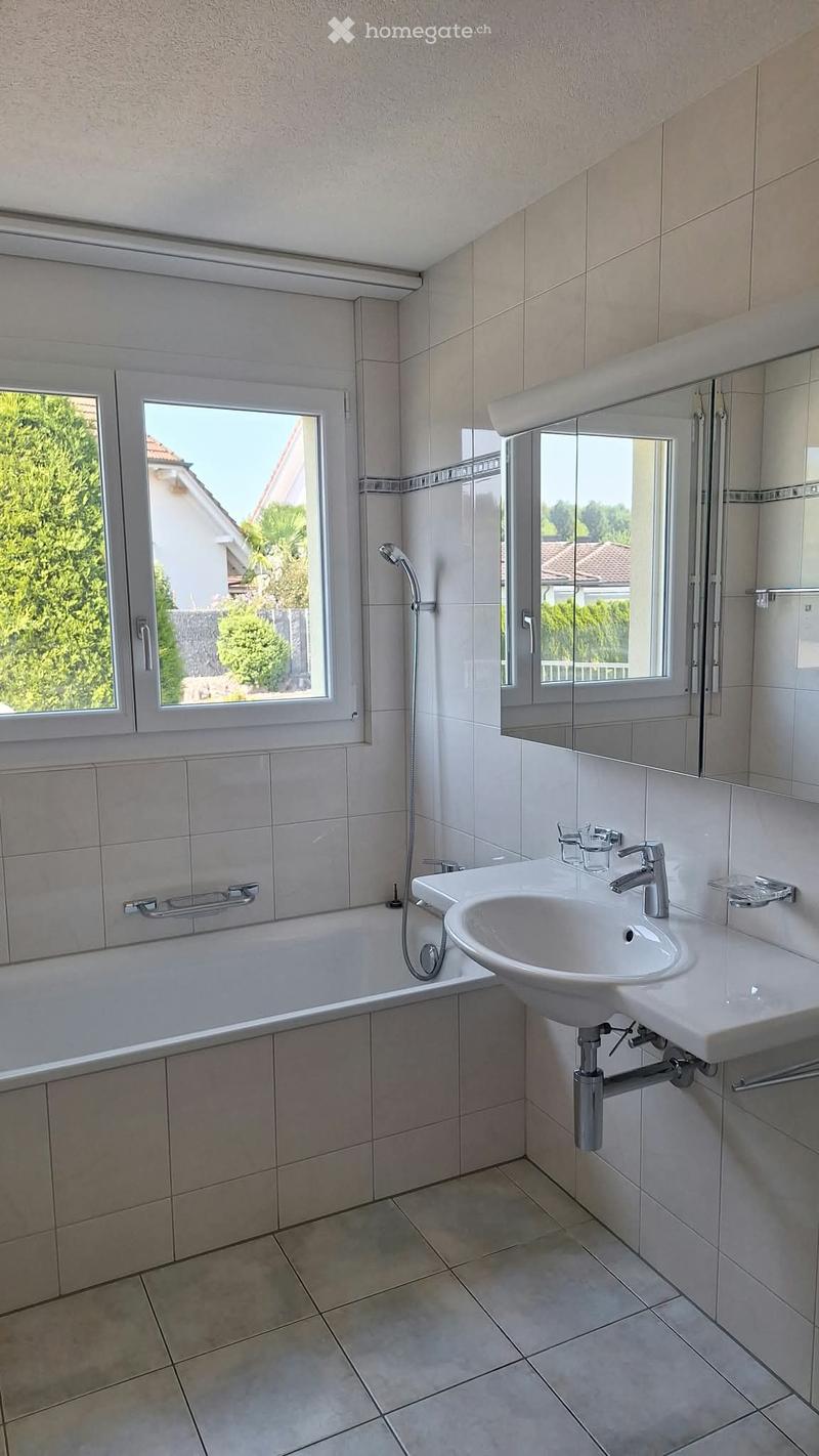 Modern bathroom with a bathtub, sink, mirror, and showerhead. The room features large windows with a view of greenery outside.