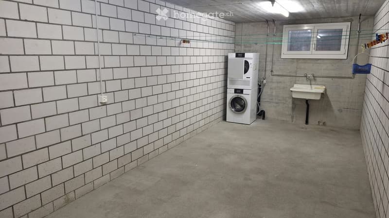 A small, tiled bathroom with a washing machine, a sink, and a window. The walls are tiled in white with black grout, and the floor is concrete.