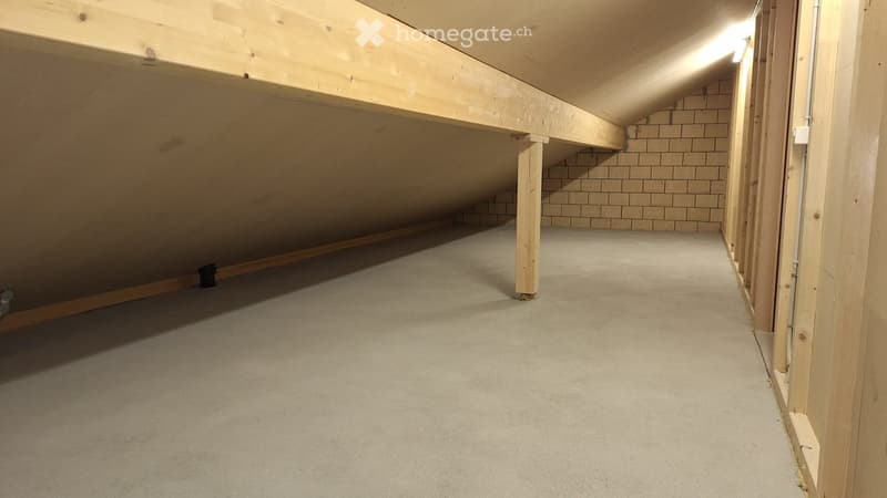Empty attic space with wooden beams, concrete floor, and exposed brick wall. No furniture or equipment present.