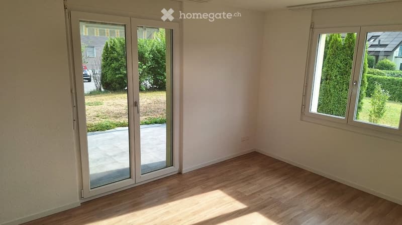 Empty room with wooden flooring, large sliding glass door leading to a patio, and a window with white frame. The walls are light-colored, and the space is well-lit with natural light.
