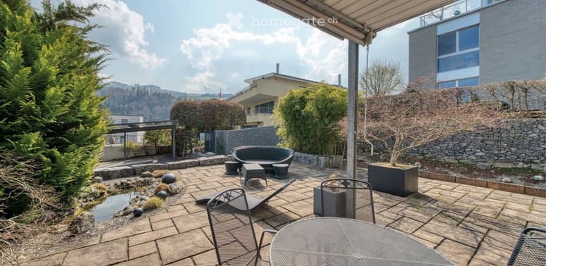 Well-maintained outdoor patio with stone tiles, seating area including a sofa, chairs, and a table, surrounded by greenery and a small water feature.