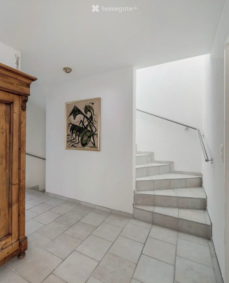 A narrow hallway with tiled flooring, white walls, a wooden cabinet on the left, and a staircase leading upwards with a metal railing on the right. A framed artwork is mounted on the wall near the staircase.
