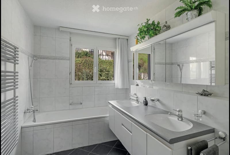 Modern bathroom with a bathtub, double sink, large mirror, and built-in cabinets. Features tiled walls, a window with white curtains, and a radiator.