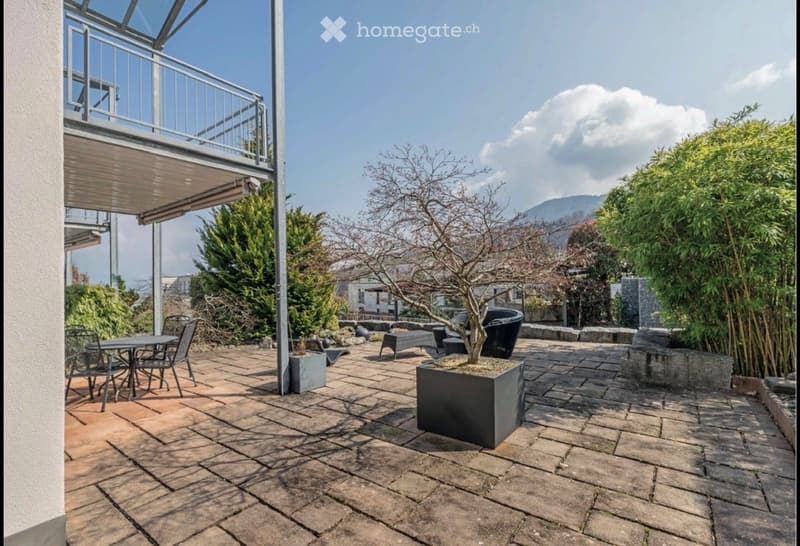 Patio with stone tiles, outdoor furniture including a table and chairs, a small tree in a planter, and a view of mountains in the background.