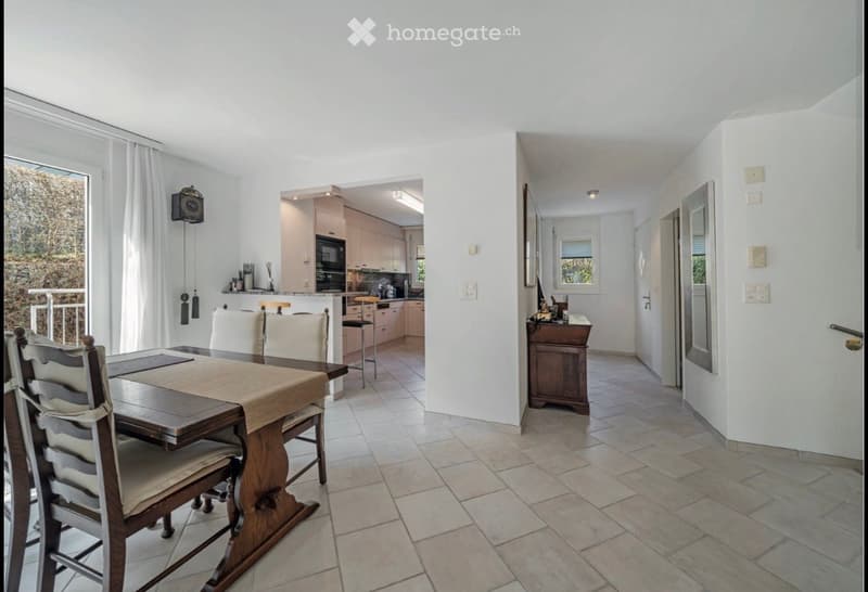 Bright, open-plan dining and kitchen area with a wooden dining table, chairs, and modern kitchen cabinets. A small hallway leads to another room.
