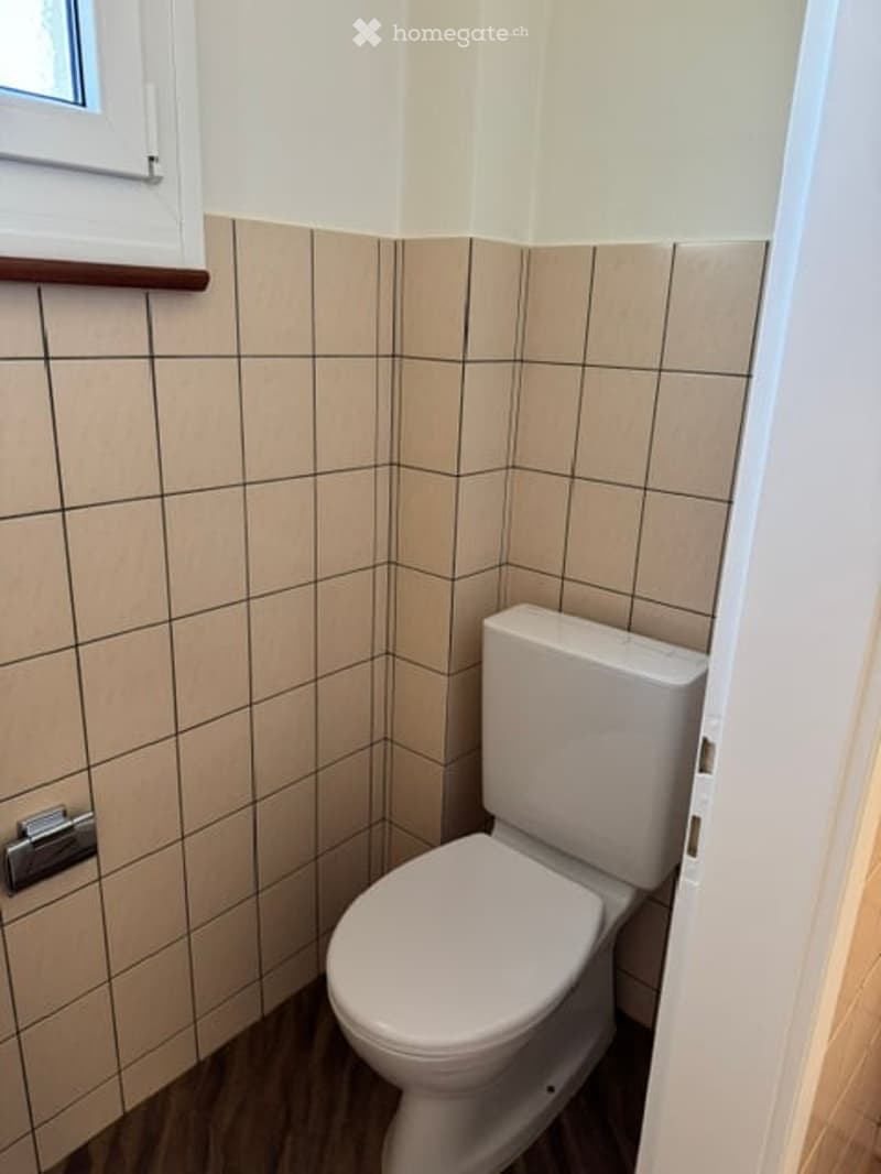 Small bathroom with beige tiled walls, wooden floor, and a white toilet. A window with a wooden sill is visible on the left side.