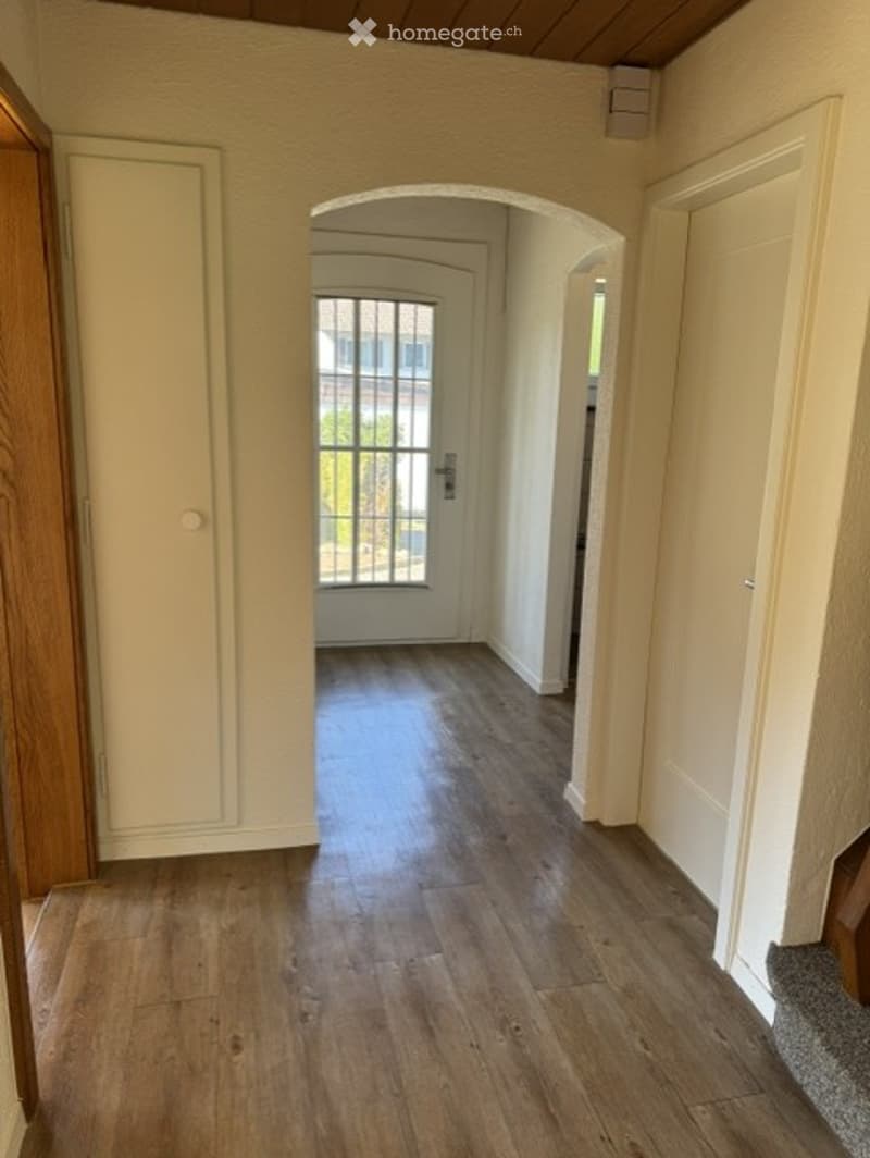 Empty hallway with wooden flooring, white walls, and an arched doorway leading to another room.