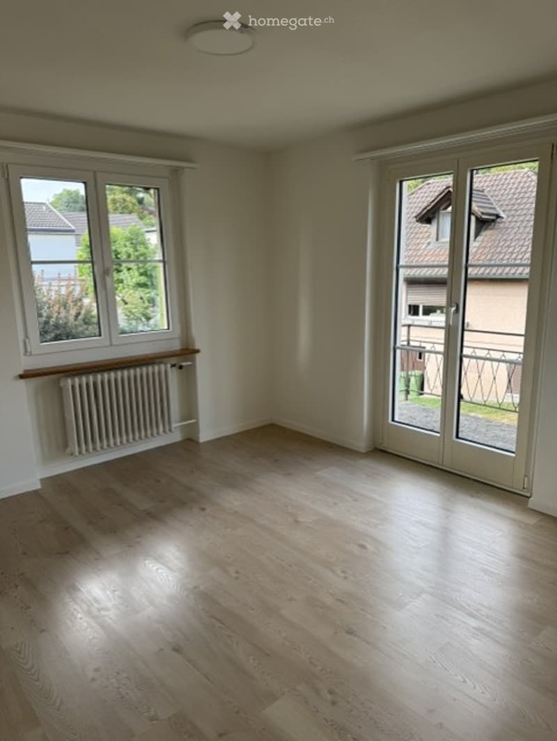 Empty room with wooden flooring, white walls, large windows, and a sliding glass door leading to a balcony.