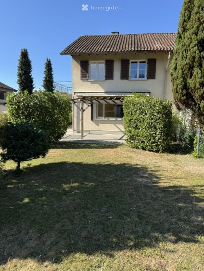 Two-story house with a tiled roof, beige walls, and brown shutters. The house is surrounded by a well-maintained lawn, trimmed bushes, and tall pine trees.