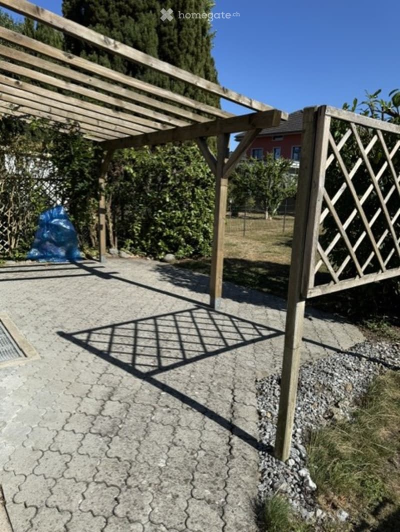 Wooden pergola with lattice fence, gravel patio, surrounding greenery, and a red house in the background.
