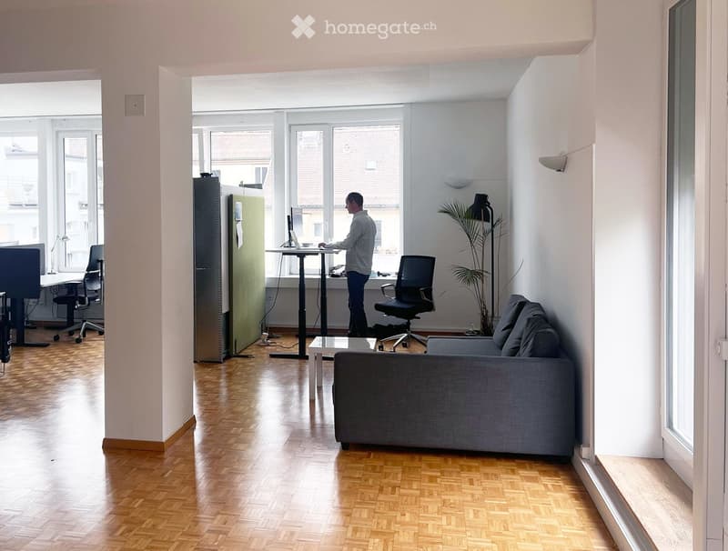 Modern office space with a wooden floor, large windows, a sofa, a chair, a standing desk with a laptop, and a plant in the corner.