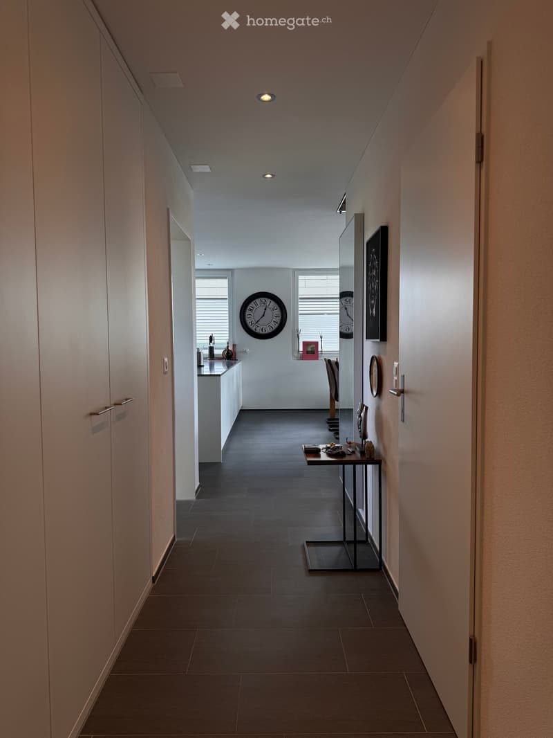 Modern hallway with dark tiled floor, white walls, built-in cabinets, and a small table with decorative items. A large wall clock is visible in the background.