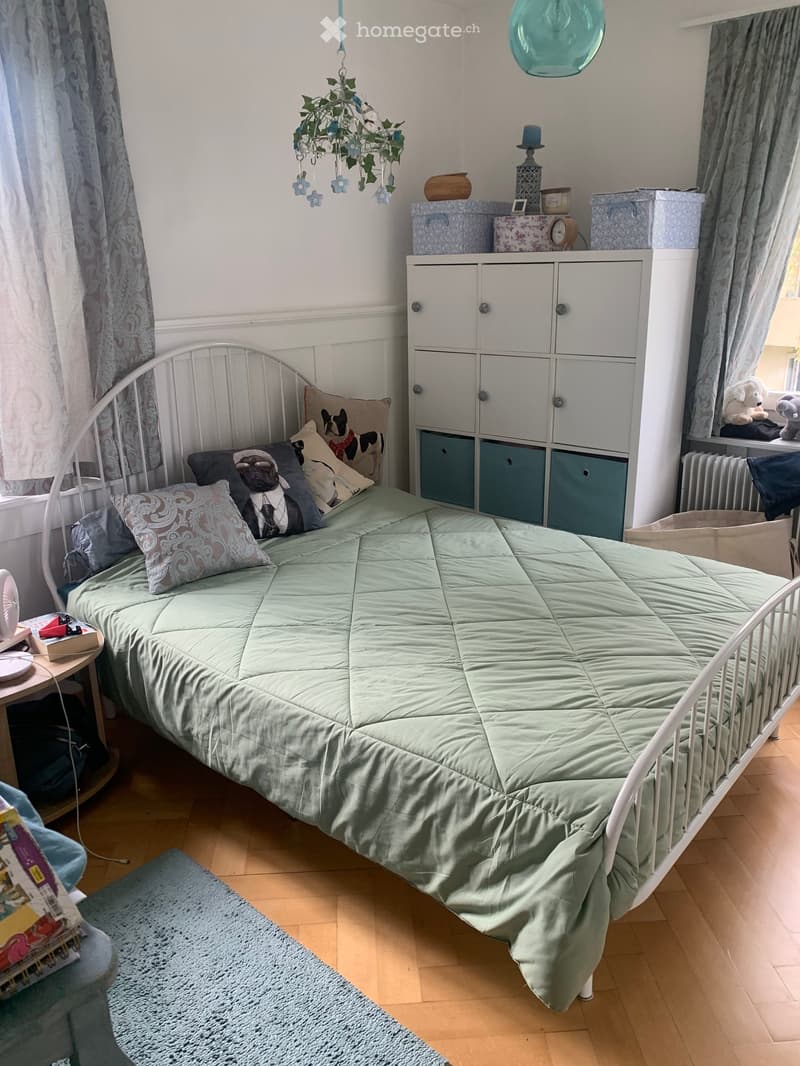 A cozy bedroom with a single bed, pillows, a blanket, a cabinet, a small table with a fan, and decorative items including a lamp and a clock. The room features wooden flooring and a window with curtains.