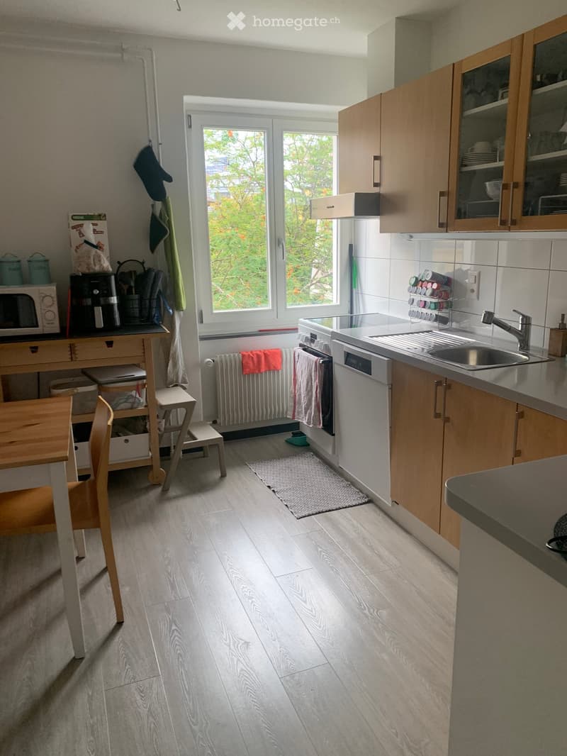 Modern kitchen with light wooden cabinets, stainless steel sink, dishwasher, microwave, coffee maker, and a small dining area with a table and chairs. The space features a large window with a view of greenery outside.