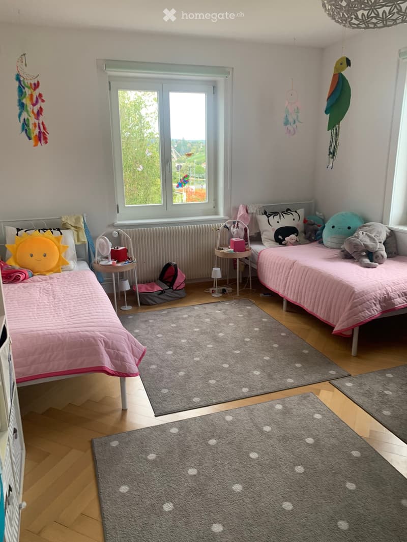 Cozy children's bedroom with two single beds, pink bedding, nightstands, toys, and a large window with a view of greenery.