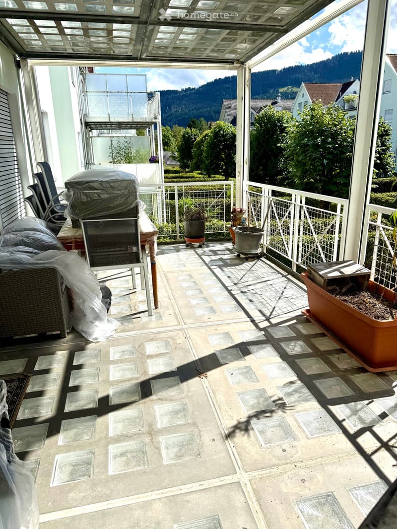 Balcony with a view of a green landscape and mountains, featuring a wicker sofa, a table, and potted plants.