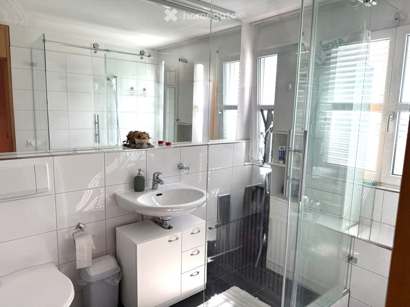 Modern bathroom with white tiles, a sink with a modern faucet, a large mirror, a glass-enclosed shower, a toilet, and a small cabinet with drawers.