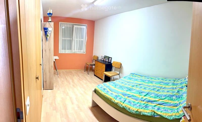 Cozy bedroom with wooden flooring, a single bed with a blue and green striped duvet, a small table, a chair, and a cabinet with picture frames on top.