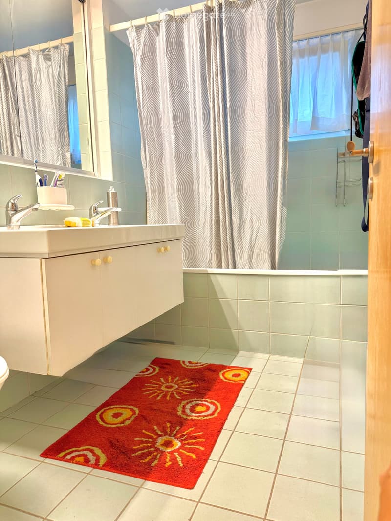 Bathroom with a white sink, two faucets, a toothbrush holder, a bathtub with a shower curtain, and a red bathroom rug with sun-shaped designs.