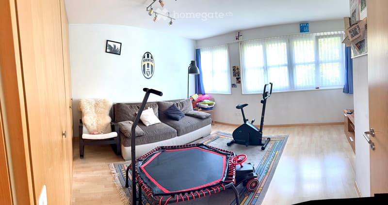 Living room with a grey sofa, a white armchair, an exercise bike, a small trampoline, a side table, and a floor lamp. The room has large windows with curtains and a light wooden floor.