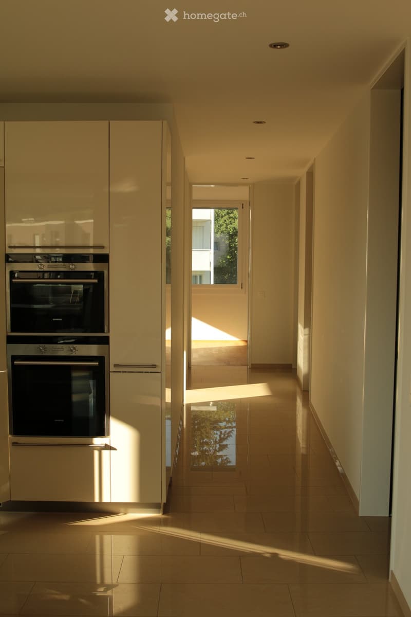 Modern hallway with light-colored walls, tiled floor, and built-in cabinets with stainless steel appliances visible on the left side. Sunlight streams in from an open doorway at the end of the corridor.