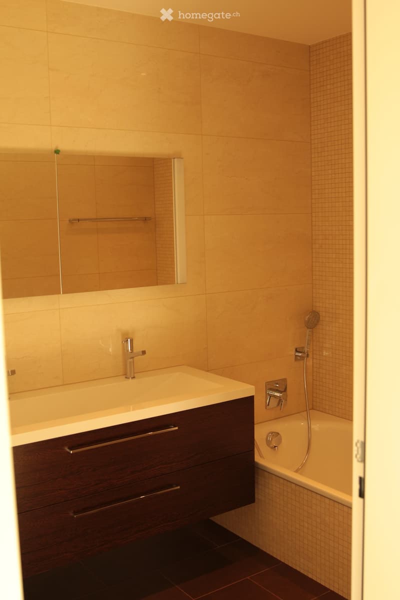 Modern bathroom with a dark wooden vanity, white sink, chrome faucet, bathtub, and showerhead. Light-colored tiles and a large mirror complete the space.