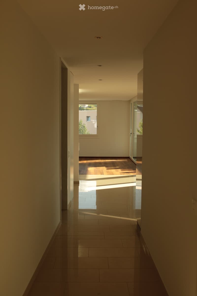 Modern hallway with tiled floor, recessed lighting, and a window at the end, leading to another room with wooden flooring.