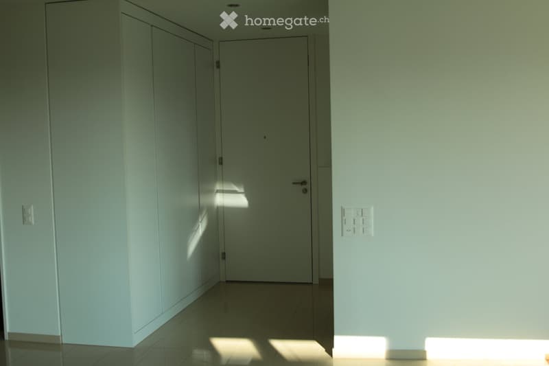 A clean, modern hallway with light green walls, tiled floor, and a closed door at the end. The space is well-lit with natural light casting shadows on the floor.