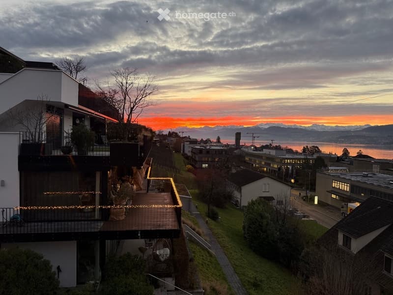 Modern multi-story building with balconies and outdoor seating areas, overlooking a scenic waterfront at sunset with mountains in the distance.