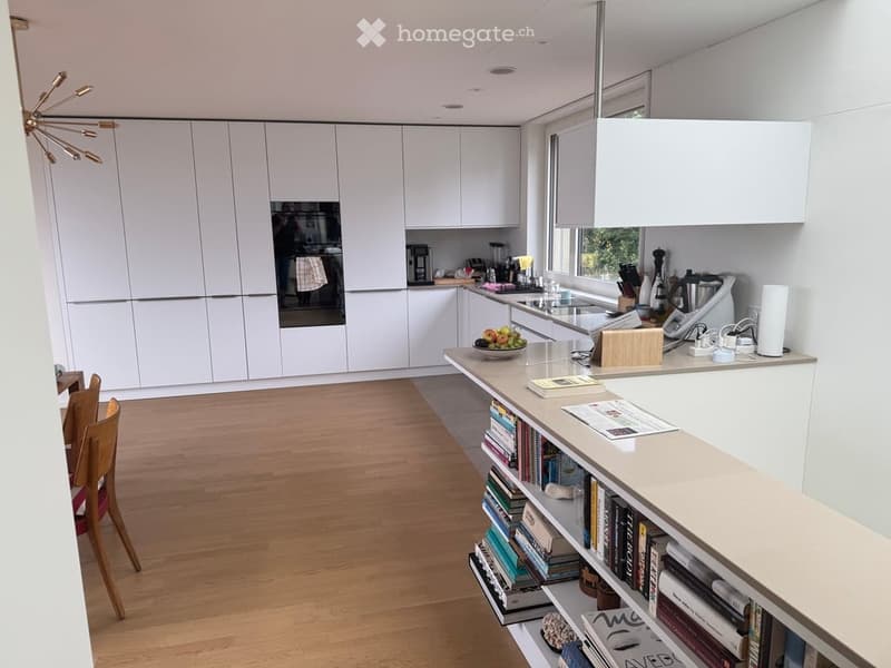 Modern kitchen with white cabinetry, a central island, and a variety of appliances including a sink, oven, and coffee maker. The island features a countertop with a bowl of fruit, a lamp, and a stack of books. Wooden chairs and a small table are visible i