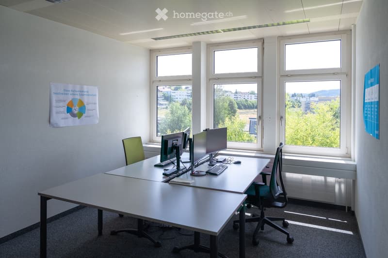 Bright office space with large windows, two desks, two chairs, two computer monitors, a keyboard, and a mouse. Posters on the walls.