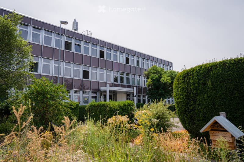 Modern multi-story building with large windows and a sleek design, surrounded by well-maintained gardens and greenery.