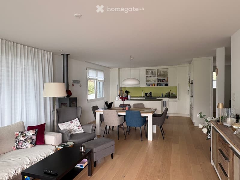 Open-plan living and dining area with light wooden flooring, a grey sofa with floral cushions, a coffee table, an armchair, a dining table with chairs, a laptop, and a modern kitchen with white cabinets and a green backsplash.