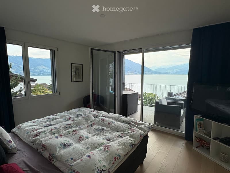 Bright bedroom with wooden flooring, large windows, and a scenic mountain view. Features a bed with a floral duvet, a chair, a small table, and a shelf with books and decorative items.