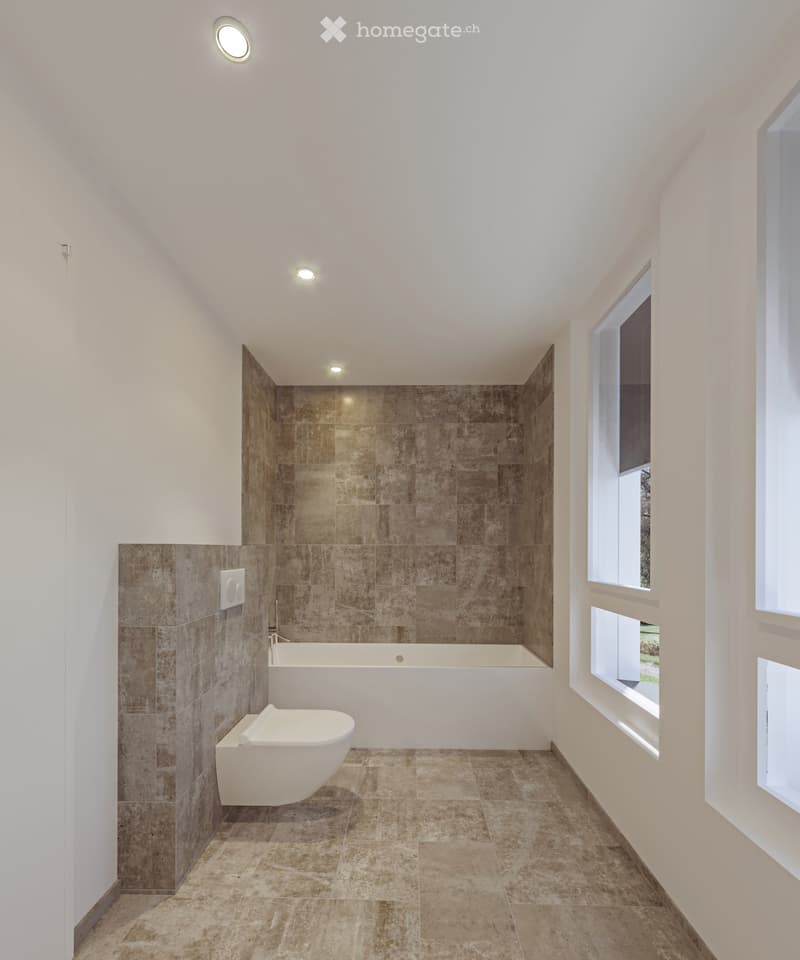 Modern bathroom with a bathtub, toilet, and large windows. The walls are tiled in a warm, earthy tone, and the floor is made of large, light brown tiles.