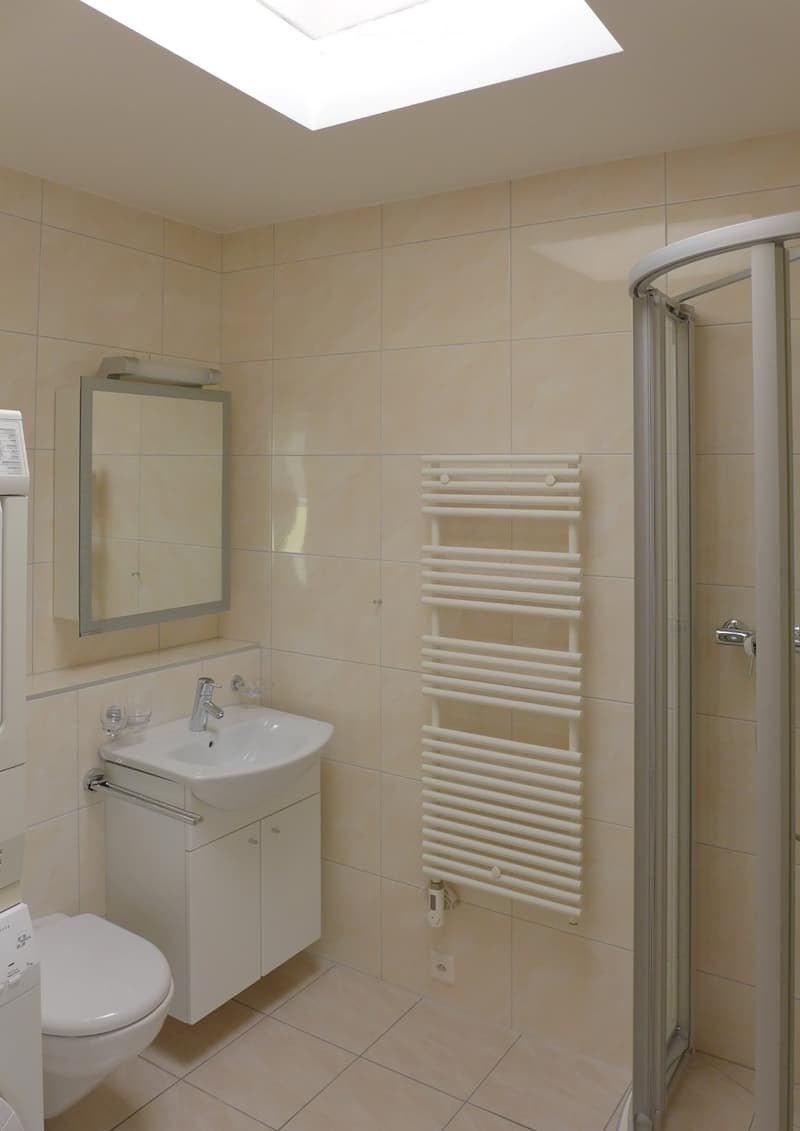 Modern bathroom with beige tiles, a white sink with a mirror above it, a radiator, a toilet, and a glass-enclosed shower area.