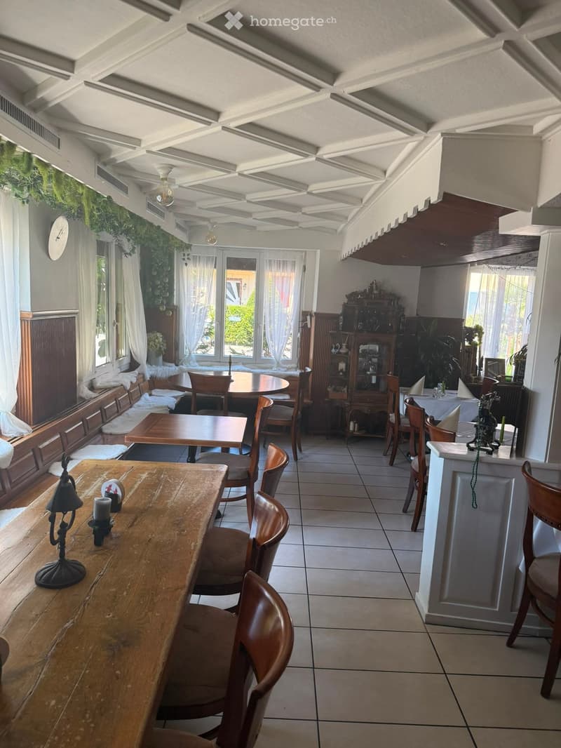 A well-lit dining area with wooden tables, chairs, and a cabinet, featuring a decorative lamp and candles on the table.