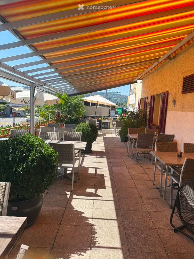 Outdoor seating area with tables, chairs, umbrellas, and potted plants under a covered walkway.