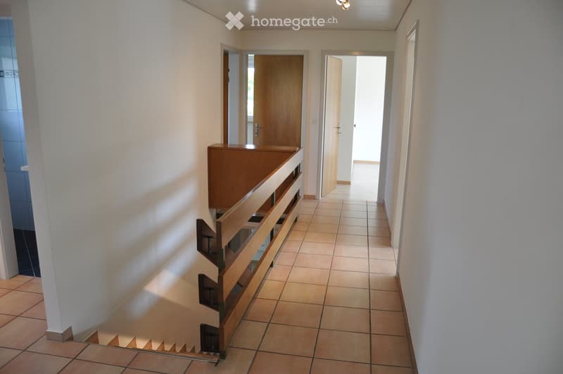 A well-lit hallway with tiled flooring, white walls, and wooden doors leading to other rooms. A wooden staircase with a railing is visible on the left side.