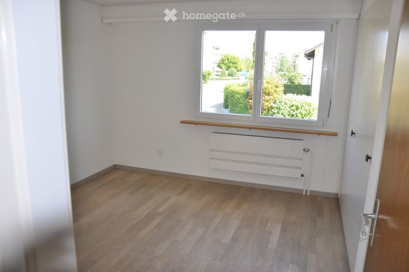 Empty room with wooden flooring, white walls, large window with wooden sill, radiator, and a door.